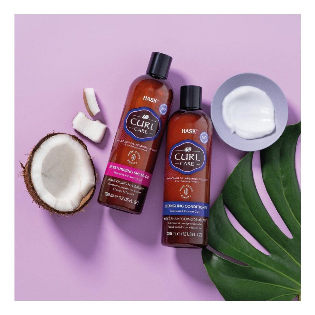 Due flaconi HASK Curl Care, uno di shampoo idratante, l'altro di balsamo districante. Pezzi di cocco e una ciotola di crema bianca sono posizionati accanto.