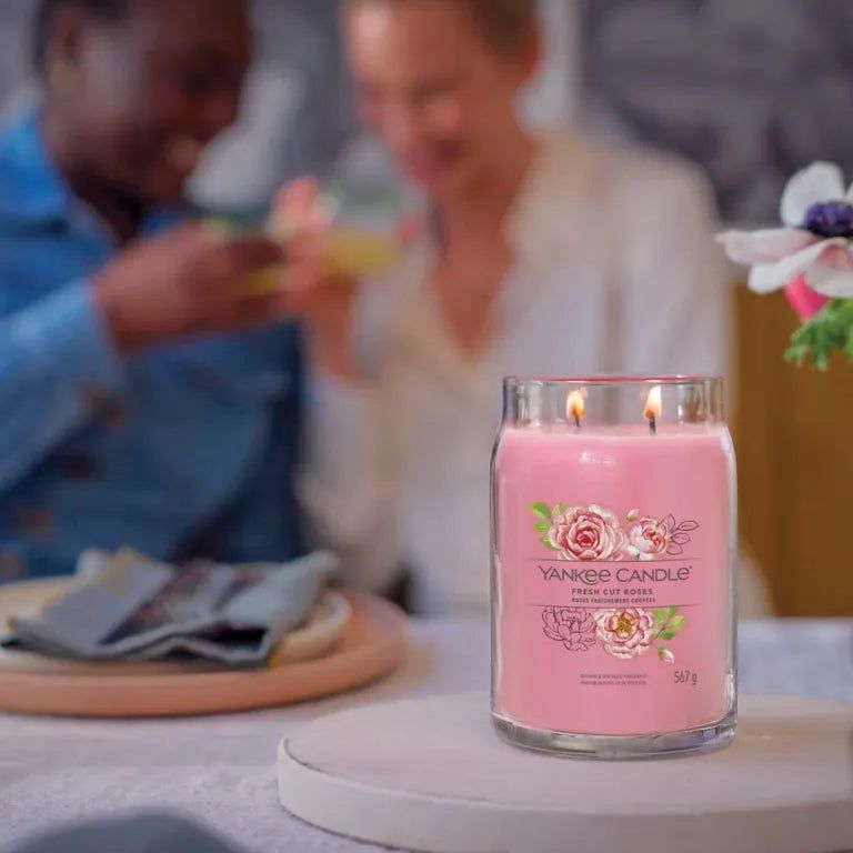 Yankee Candle accesa. Cera rosa, due fiamme. Etichetta con rose e scritta 'Fresh Cut Roses'. Su tavolo.