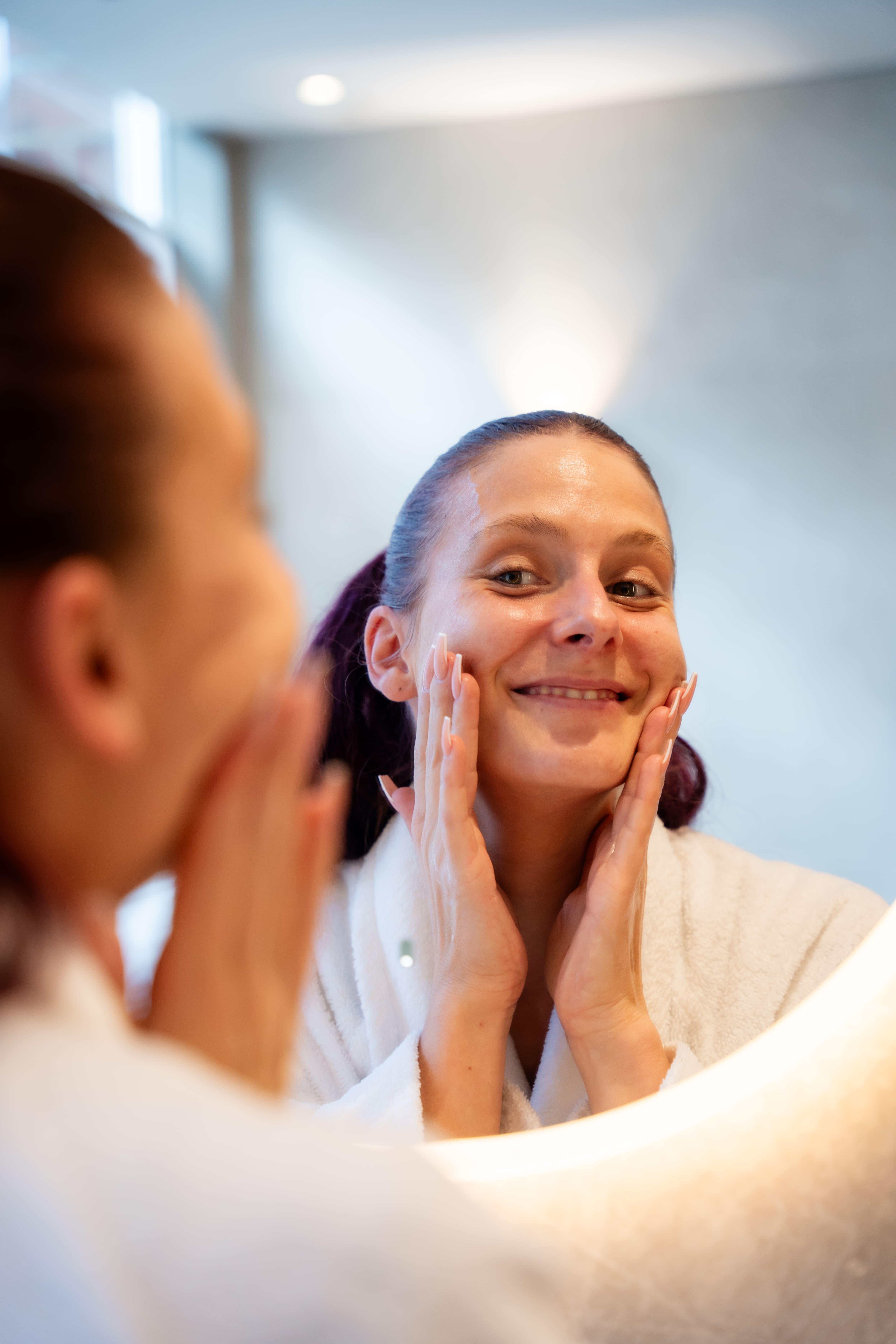 Donna in vestaglia bianca si massaggia il viso davanti a uno specchio illuminato.