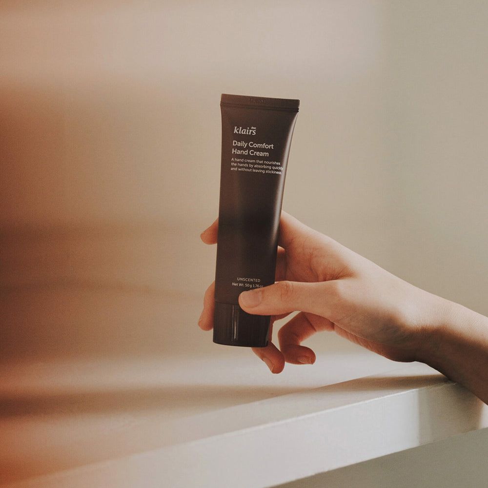 Tubo di crema per le mani marrone scuro tenuto in mano. Scritta: Klairs Daily Comfort Hand Cream.