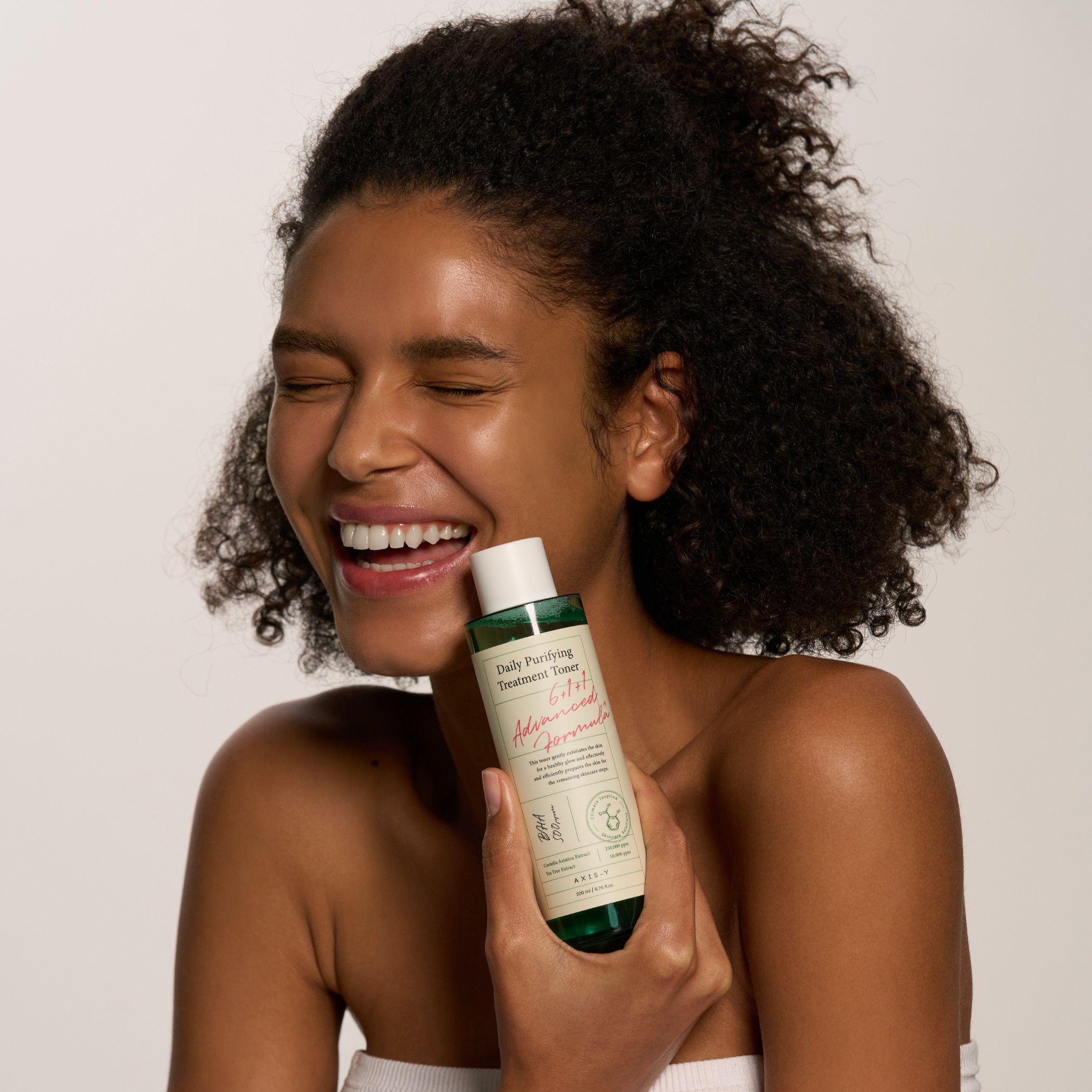 Donna sorridente tiene in mano un flacone di toner. Scritta: Daily Purifying Treatment Toner. Flacone con etichetta.