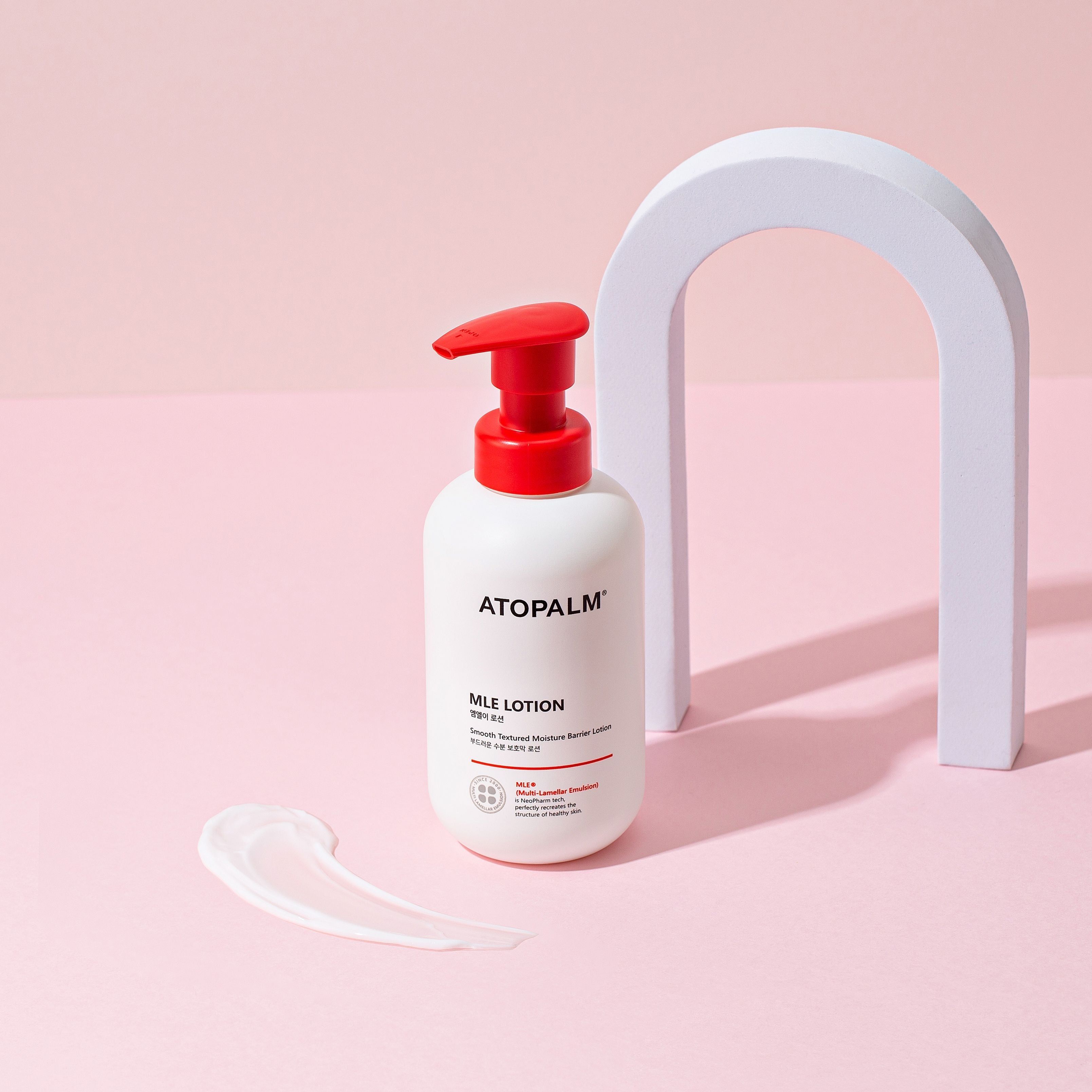 Flacone bianco con erogatore rosso. Scritta: ATOPALM MLE LOTION. Su sfondo rosa, con arco bianco e crema.