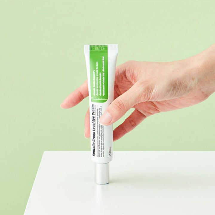 Mano che tiene un tubetto di crema per gli occhi. Tubetto bianco con tappo verde. Testo: Centella Green Level Eye Cream.
