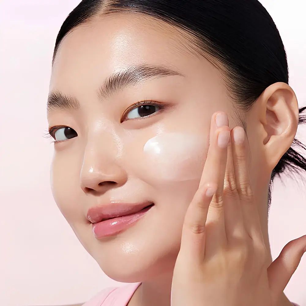 Persona applica crema sul viso. Sfondo rosa.