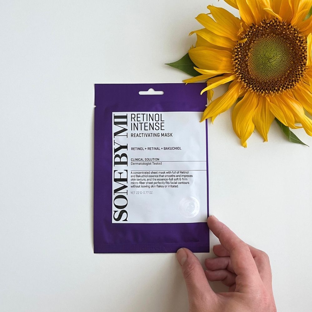 Confezione di una maschera viso con retinolo e bakuchiol, tenuta da una mano accanto a un girasole.