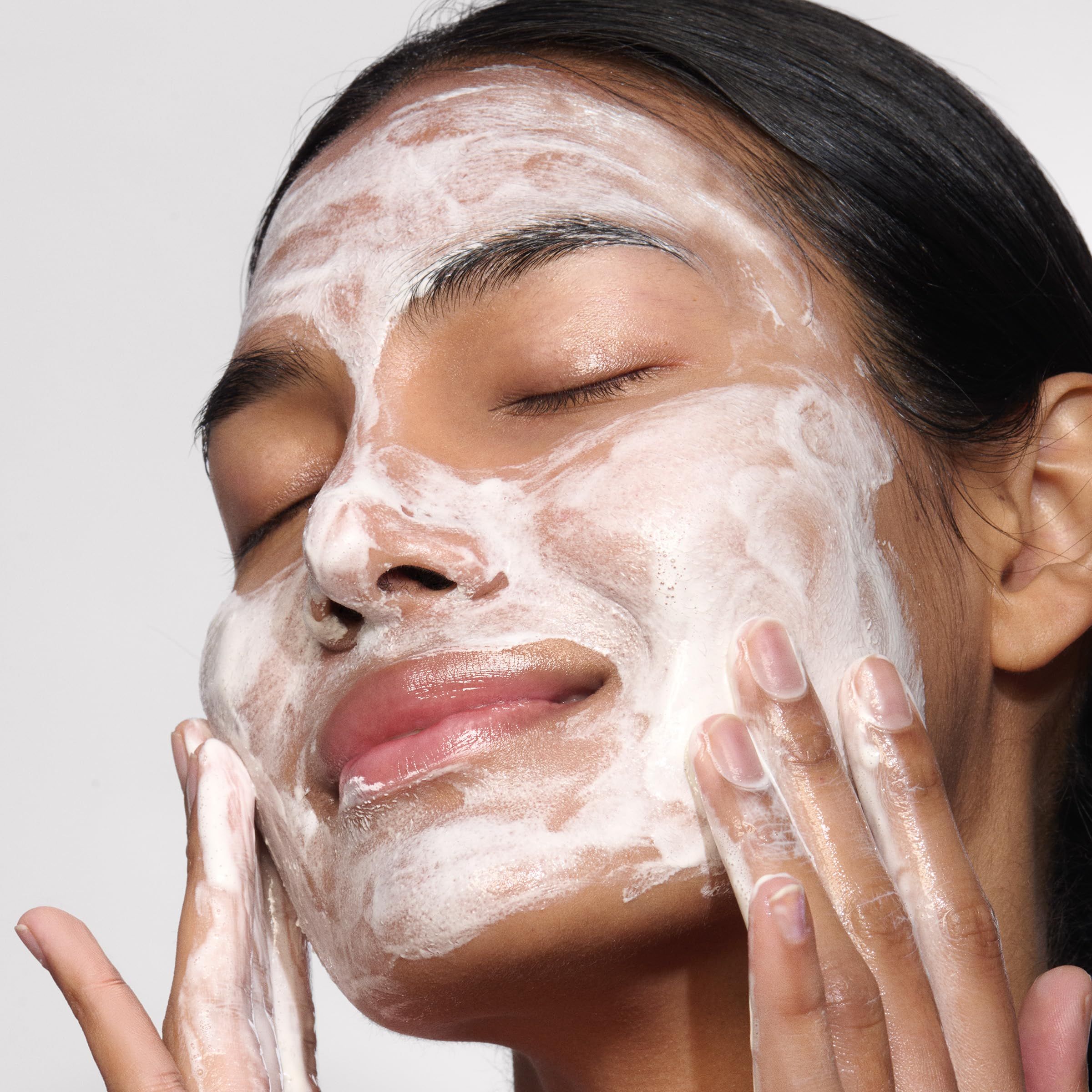 Donna con gli occhi chiusi, viso coperto di schiuma bianca. Mani che toccano il viso.