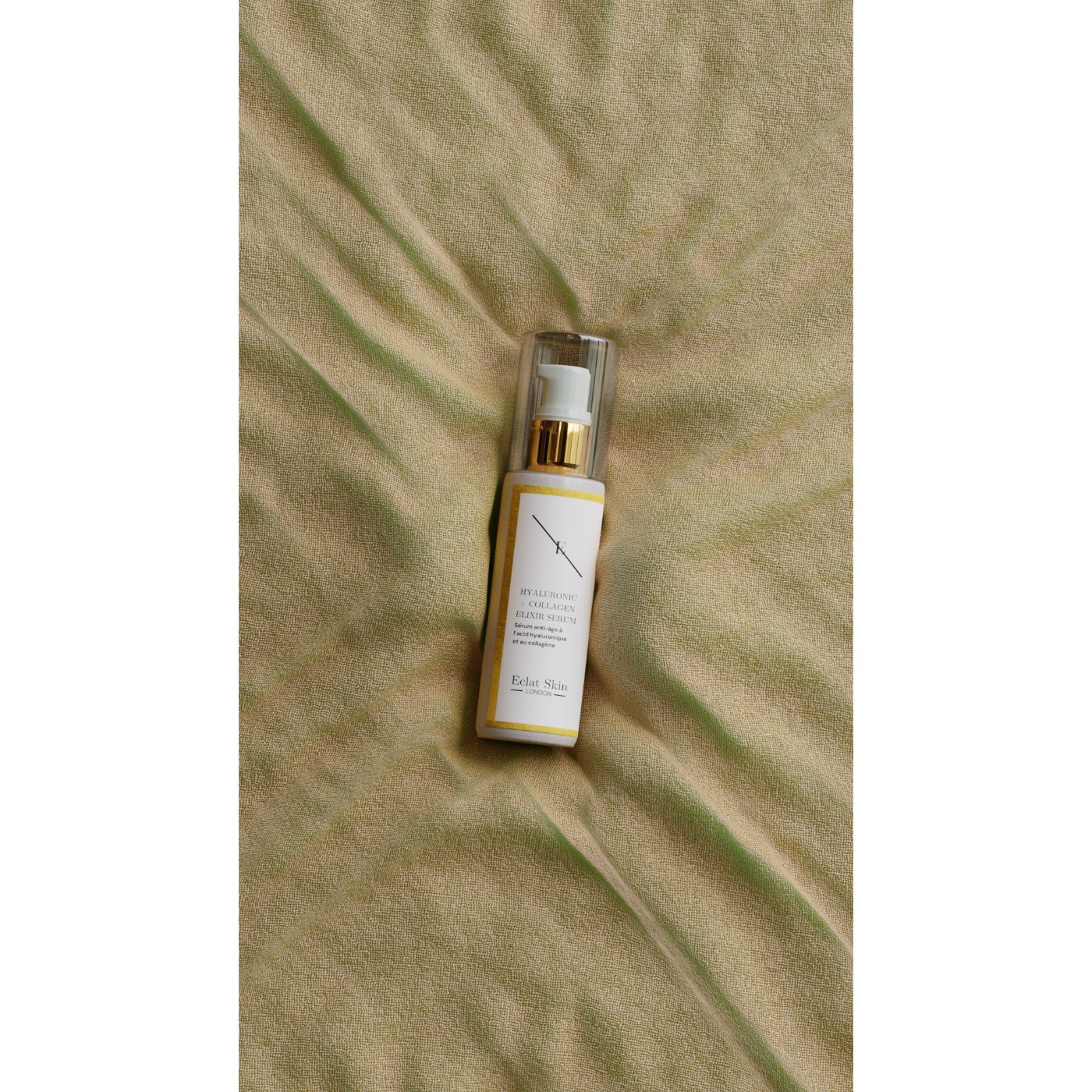 Flacone di siero con tappo dorato. Scritta: Hyaluronic Acid + Collagen Elixir Serum. Sfondo: tessuto giallo.