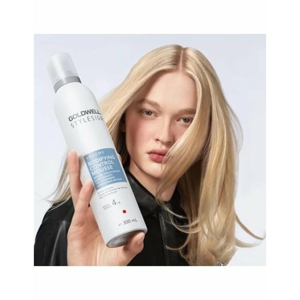 Donna tiene Goldwell Stylesign Volume Bodifying Control Mousse. Capelli biondi. Giacca nera. Etichetta del prodotto visibile.