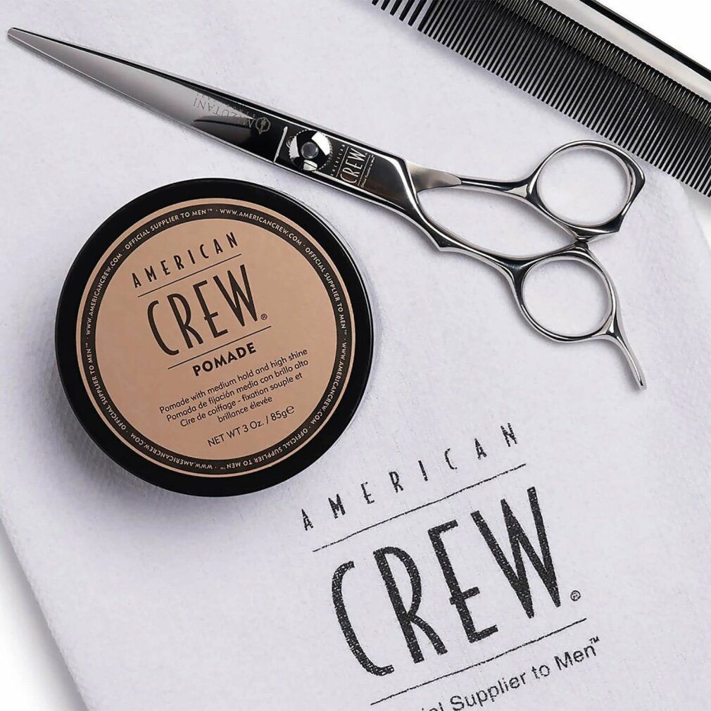 Barattolo nero con coperchio marrone. Scritta: AMERICAN CREW POMADE. Accanto, forbici, pettine e panno.