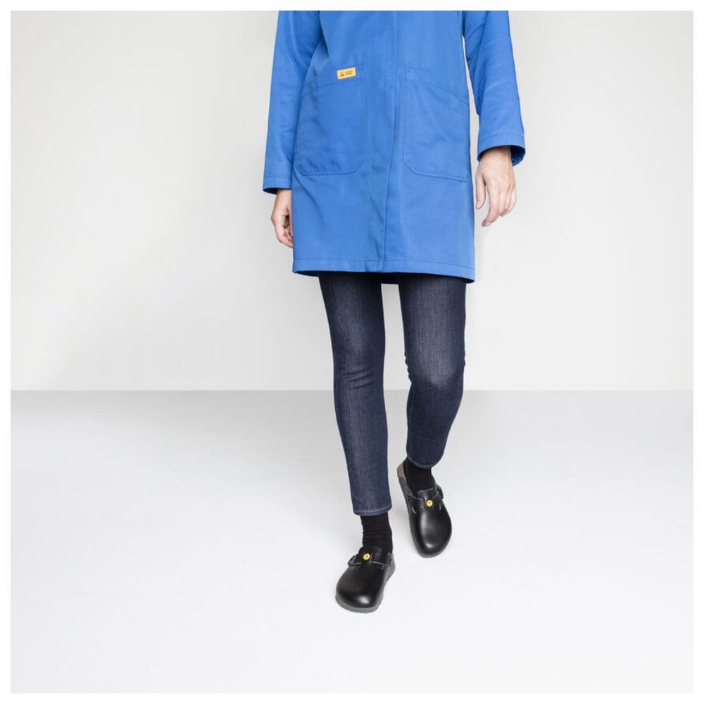 Persona che indossa zoccoli neri. Giacca da lavoro blu, jeans. Logo giallo sulle scarpe. Ambiente luminoso.