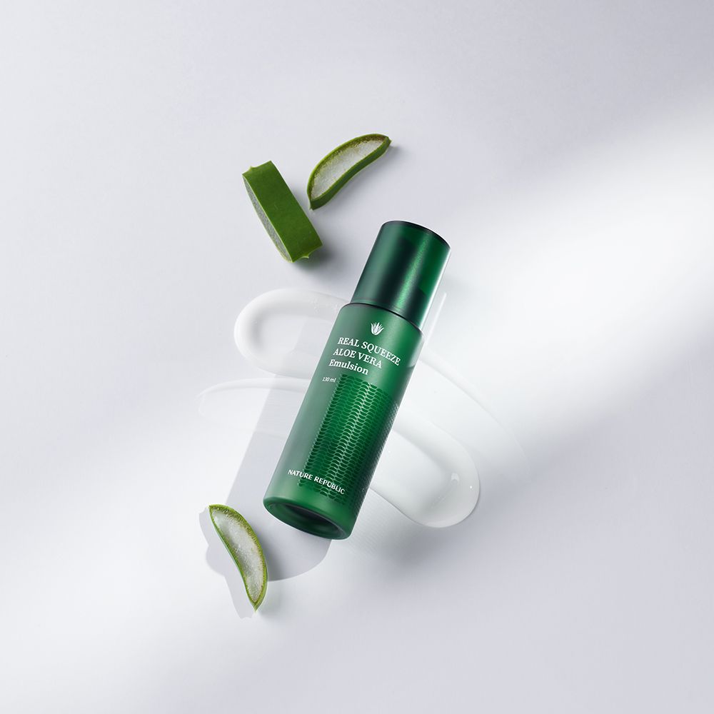 Flacone verde con testo del prodotto, circondato da pezzi di aloe vera e crema. Testo: Real Squeeze Aloe Vera Emulsion.