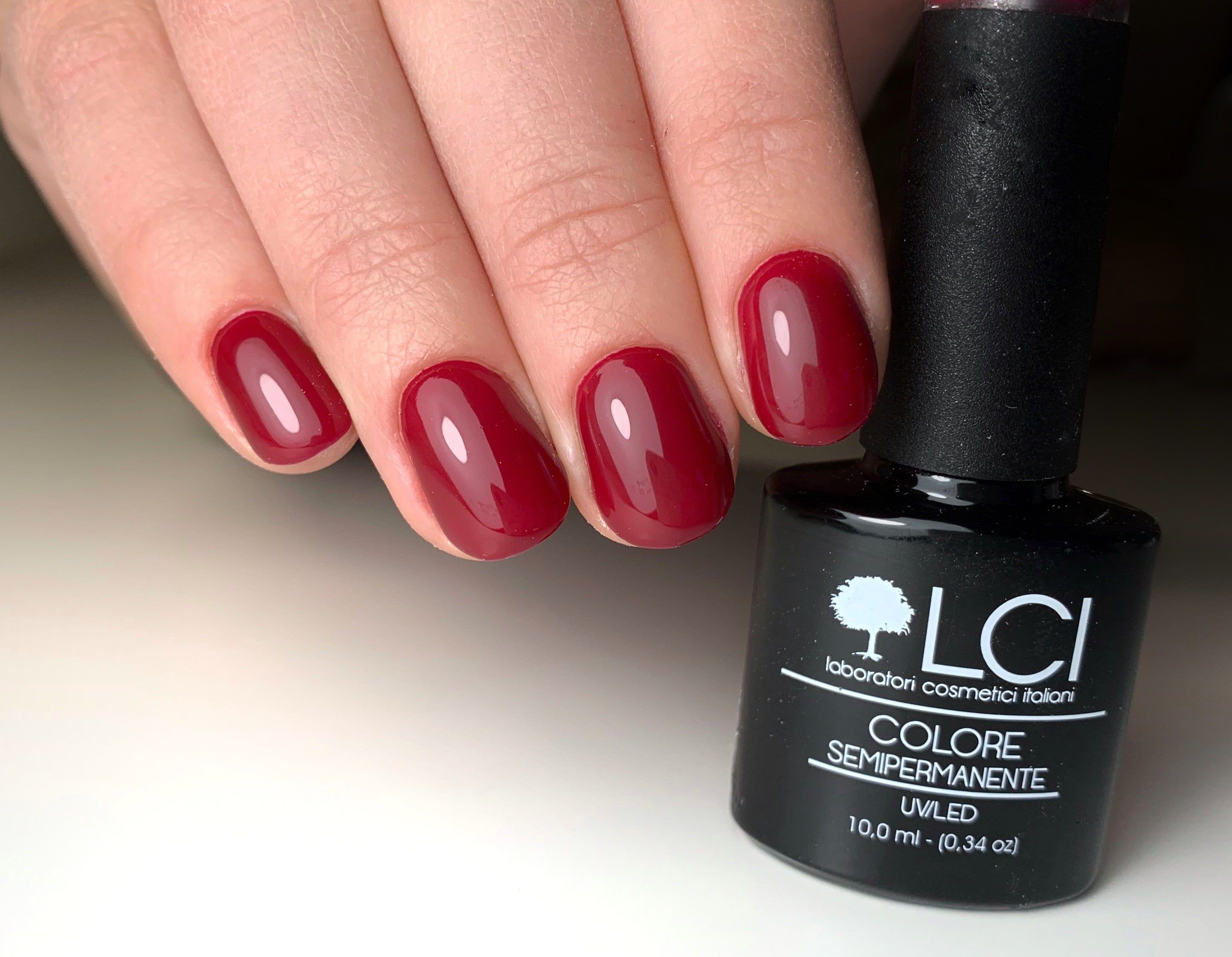 Mano con unghie laccate rosso scuro accanto a flacone di smalto. Logo LCI e scritta 'Colore Semipermanente' visibili.