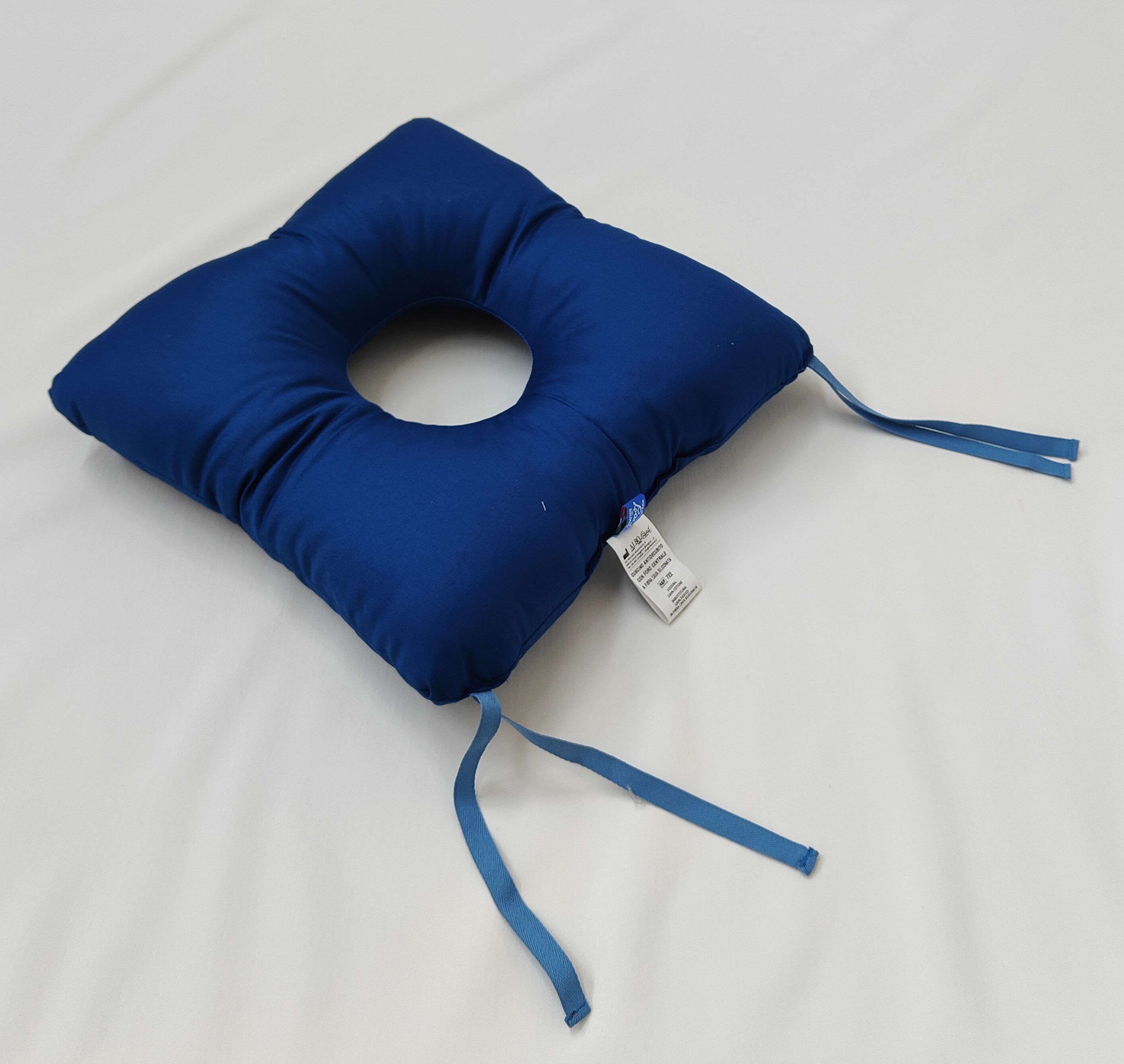 Cuscino blu quadrato con foro centrale. Nastri agli angoli. Etichetta visibile.