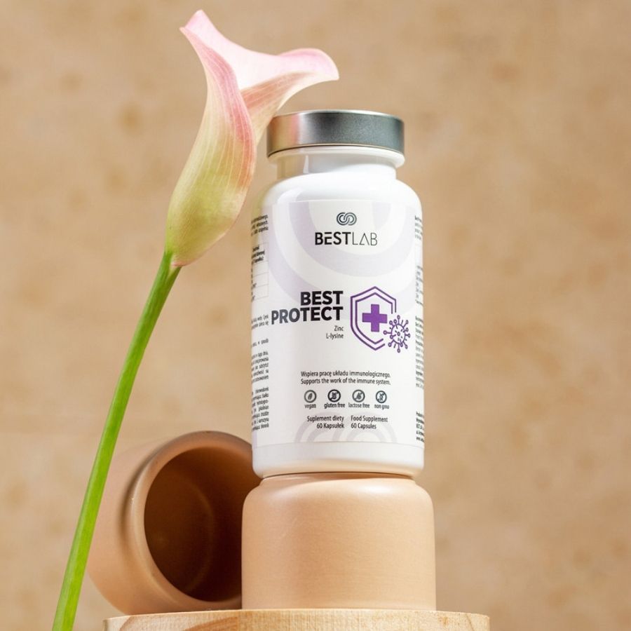 Flacone bianco con tappo argentato, posto su una base di legno. Scritta: BEST LAB, BEST PROTECT, Zinco L-Lisina. Decorazione: fiore e vaso in ceramica.