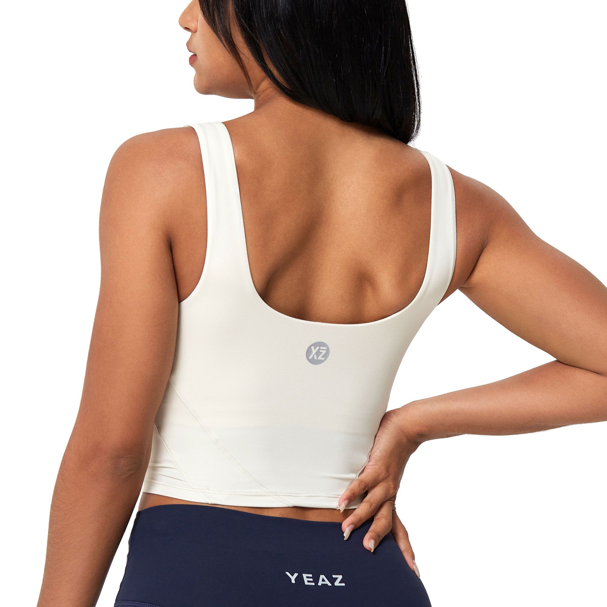 Vista posteriore di una donna con crop top color crema a scollo rotondo e piccolo logo sul retro. Leggings blu scuro con nome del marchio visibile.