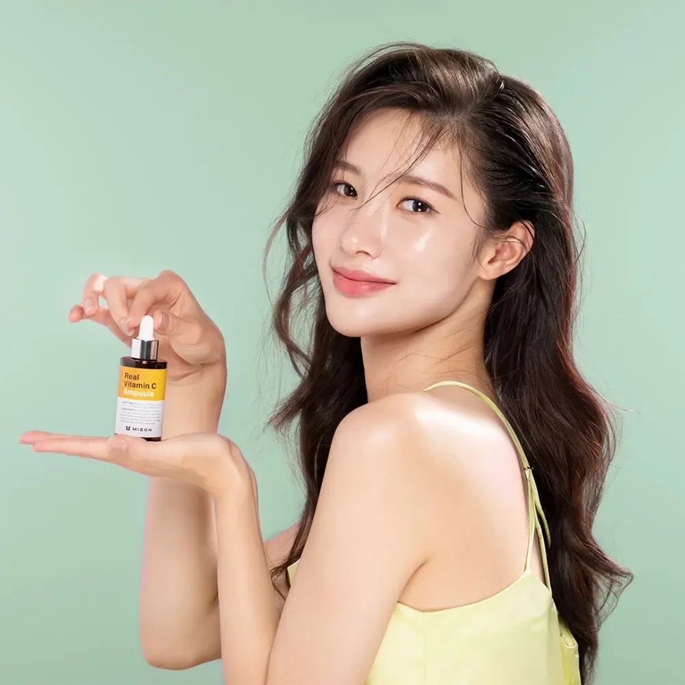 Donna con MIZON Real Vitamin C Ampoule. Etichetta gialla, flacone marrone, pipetta. Sfondo verde chiaro.