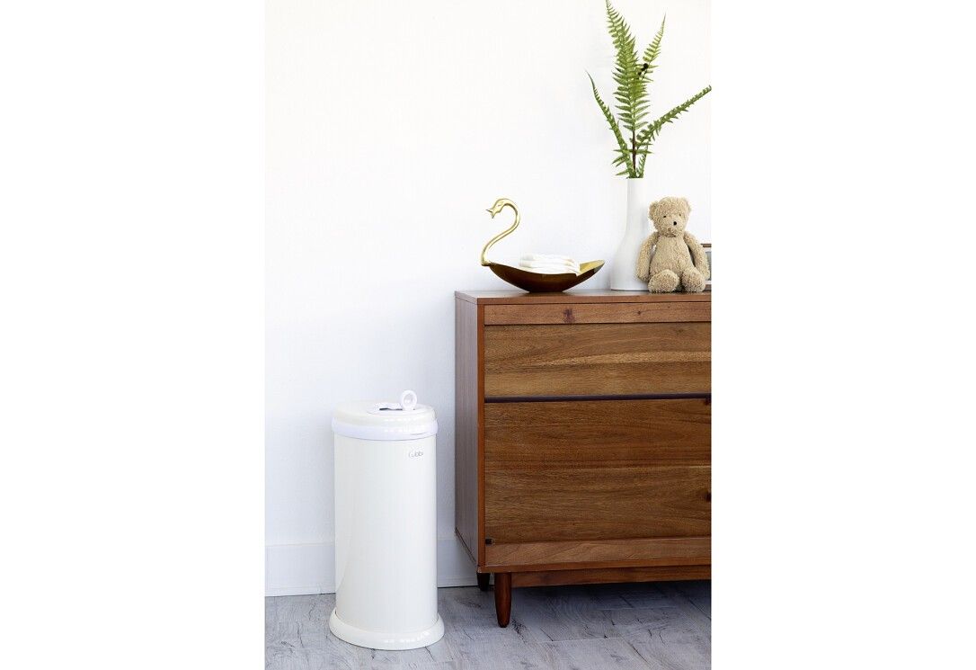 Bidone per pannolini beige. Forma cilindrica. Coperchio bianco con maniglia. Vicino a cassettiera.