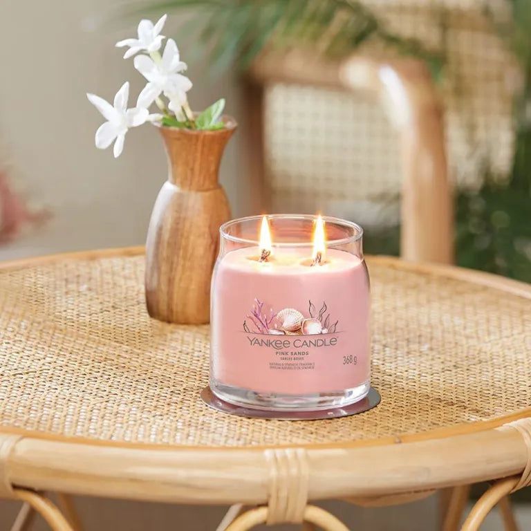 Candela rosa Yankee Candle Pink Sands su un tavolo. Due stoppini accesi. Vaso con fiori sullo sfondo.