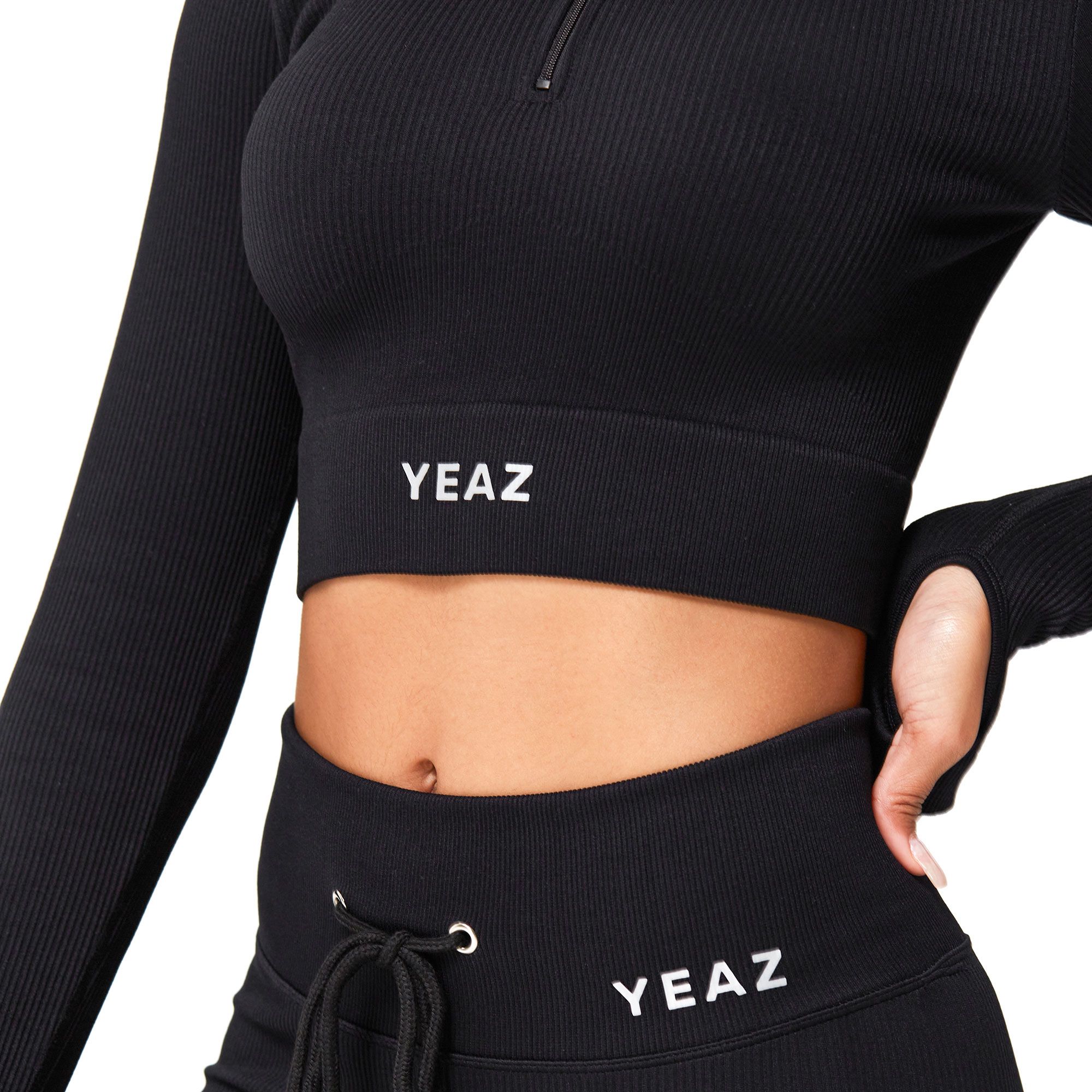 Completo sportivo nero a coste: crop top a manica lunga con zip e pantaloni coordinati con coulisse. Marchio 'YEAZ' visibile.
