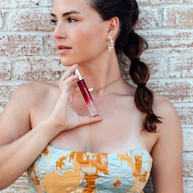 Flacone trasparente con liquido rosso. Tappo argentato. Scritta MATCHMAKER. Donna che tiene il flacone. Sfondo: muro di mattoni.