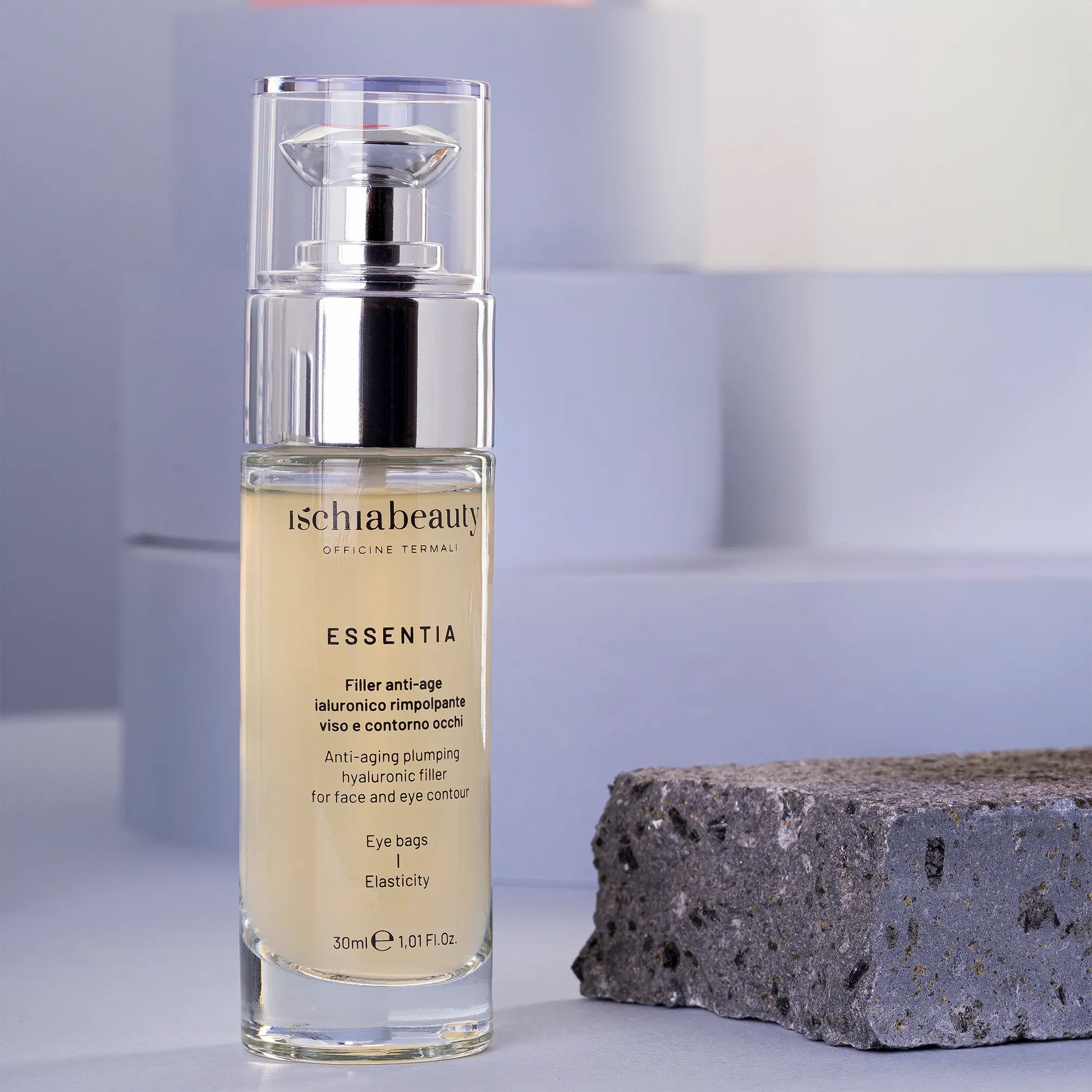Flacone in vetro con pompa argentata. Scritte: ischiabeauty, ESSENTIA, Filler anti-age, borse occhi, elasticità. Su pietra grigia.