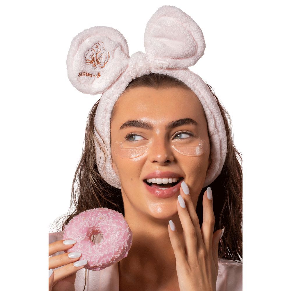 Donna con cuscinetti per occhi e fascia rosa con orecchie. Tiene in mano una ciambella rosa. Cuscinetti per occhi visibili.