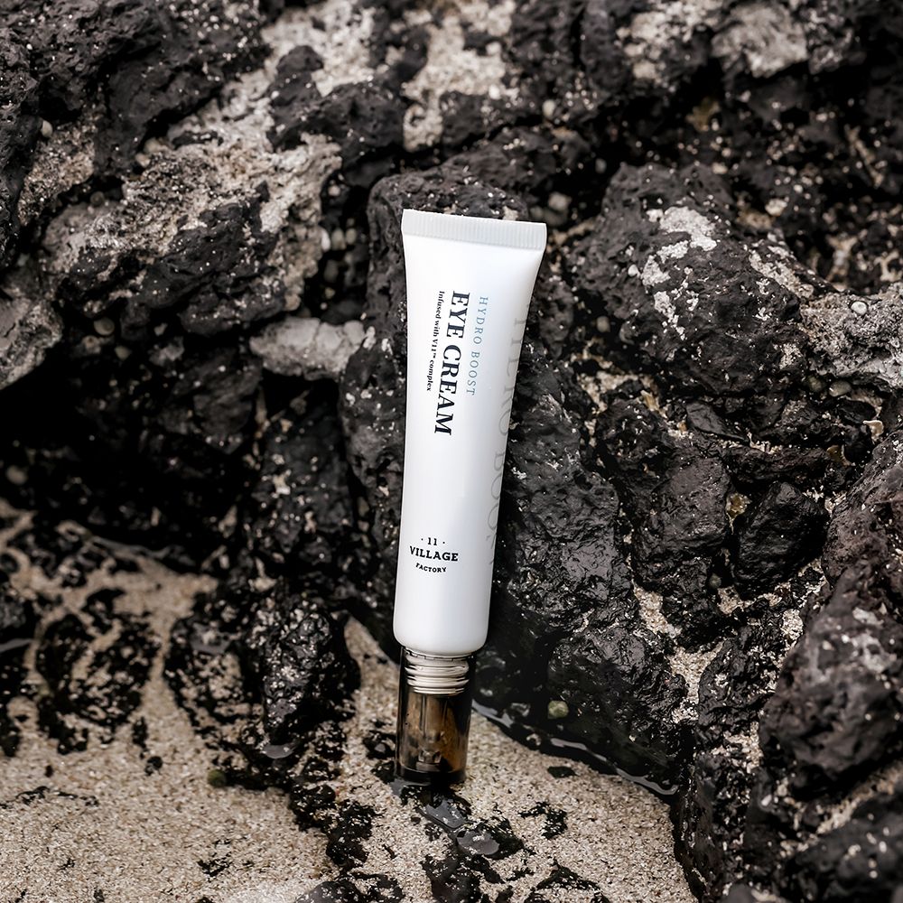 Tubo bianco su pietre scure. Scritta nera: EYE CREAM, HYDRO BOOST, VILLAGE 11 FACTORY. Chiusura marrone.