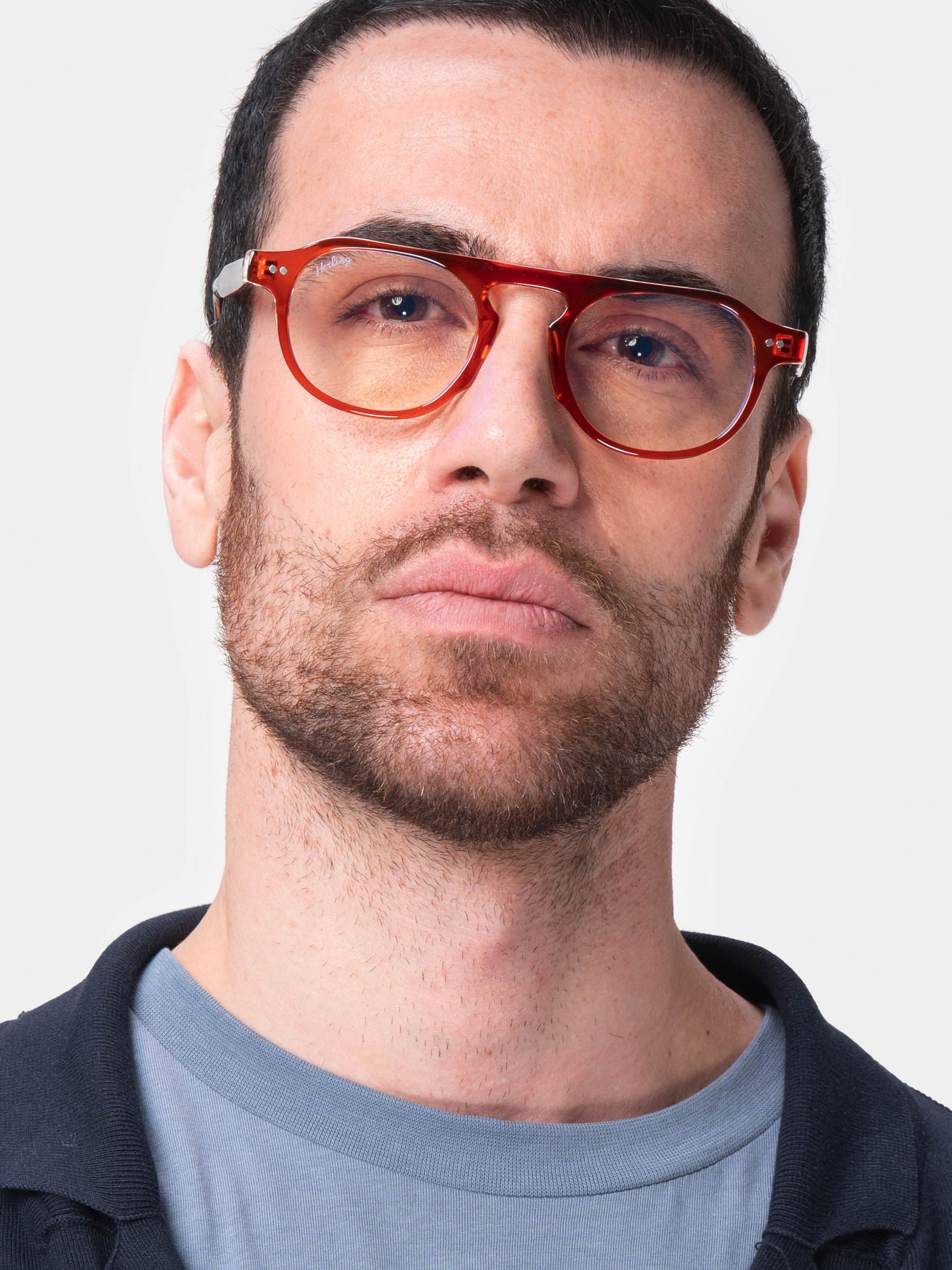 Uomo con occhiali rossi. Lenti rotonde, aste in metallo. L'uomo indossa una maglietta blu.