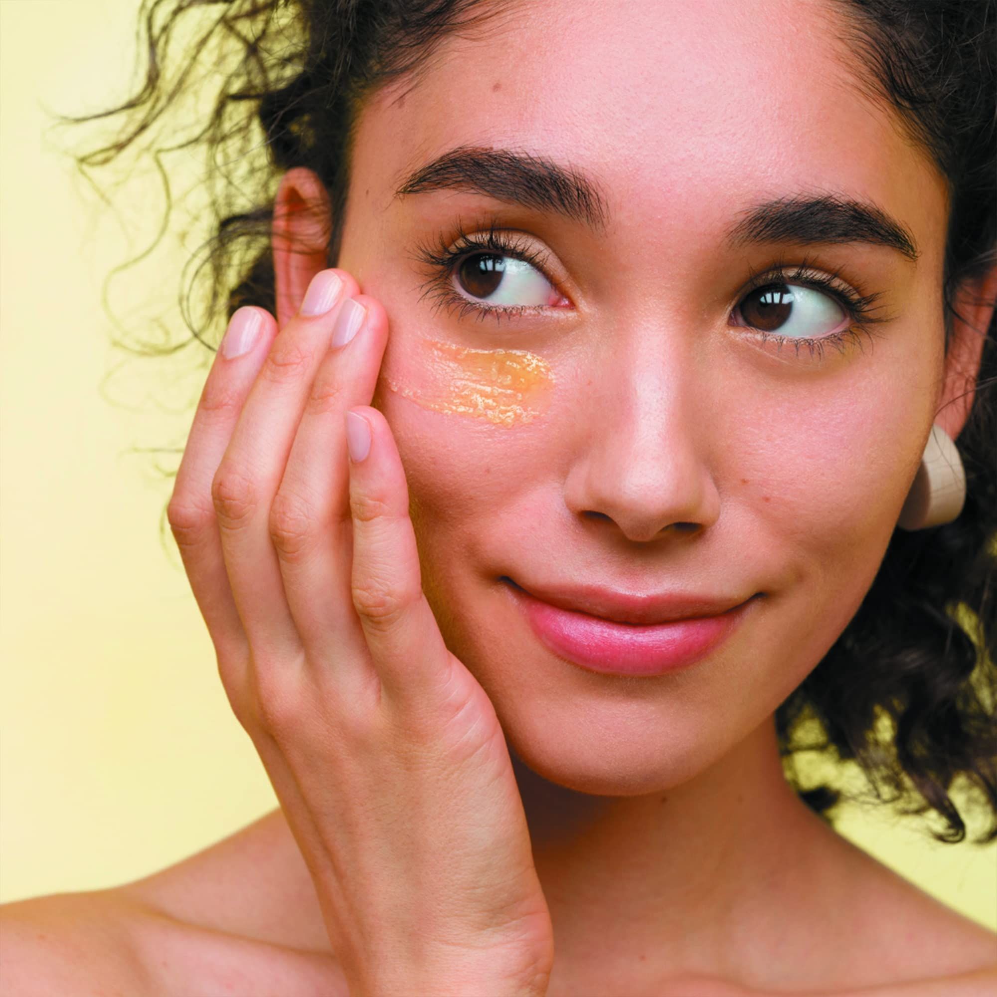 Donna applica crema giallastra sotto l'occhio. Sfondo giallo.