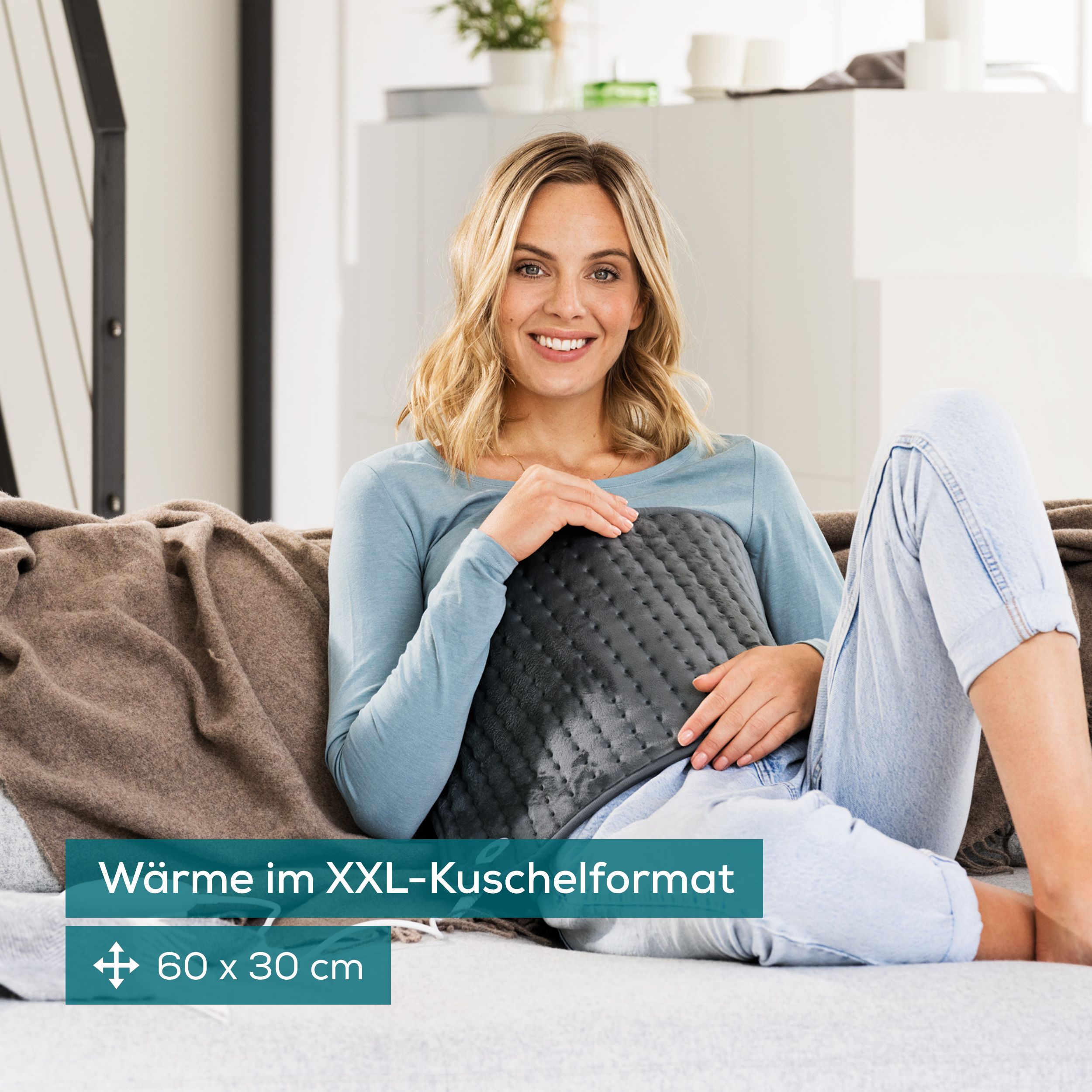 Donna seduta sul divano con cuscino riscaldante. Cuscino grigio con controllo. Testo: Calore in formato XXL. Dimensioni: 60 x 30 cm.