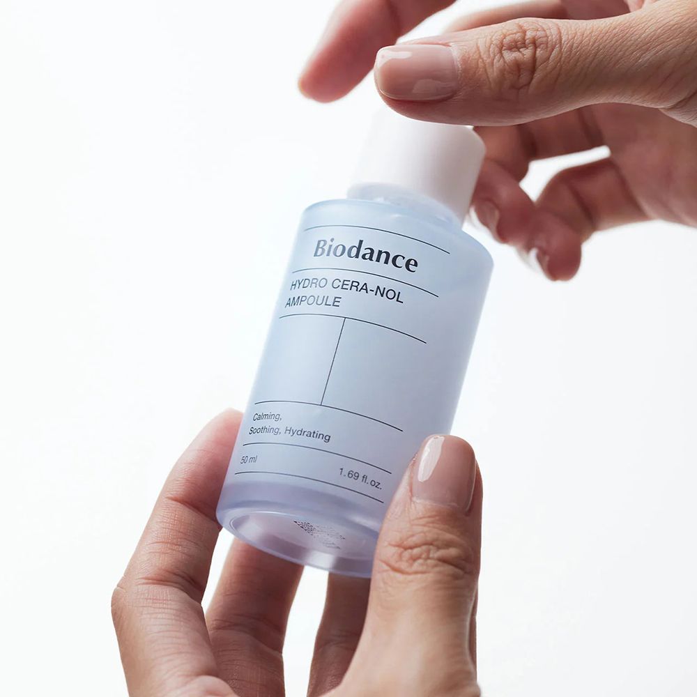 Mani che tengono un flacone blu trasparente con tappo bianco. Scritta: Biodance, HYDRO CERA-NOL, AMPULE. Ambiente luminoso.