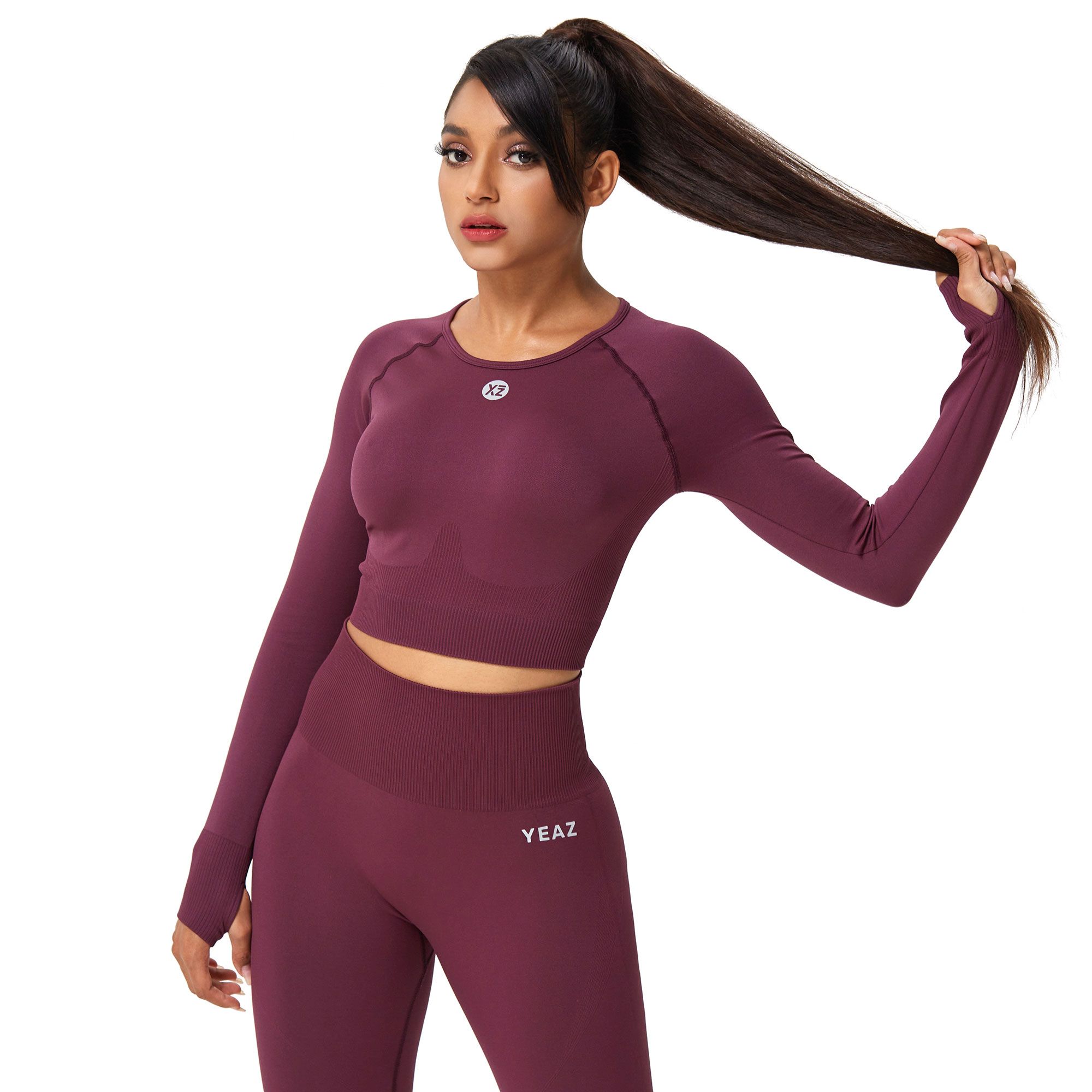 Una donna indossa un crop top bordeaux a maniche lunghe e leggings coordinati. Il top ha fori per i pollici e un piccolo logo sul petto. I leggings mostrano il nome del marchio 'YEAZ' sul fianco.
