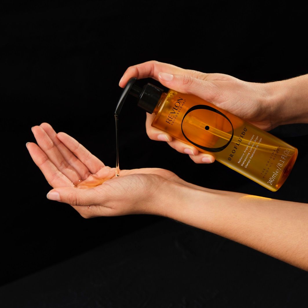 Mani che versano un liquido giallo da un flacone con erogatore nero. Scritta: Revlon Orofluido.