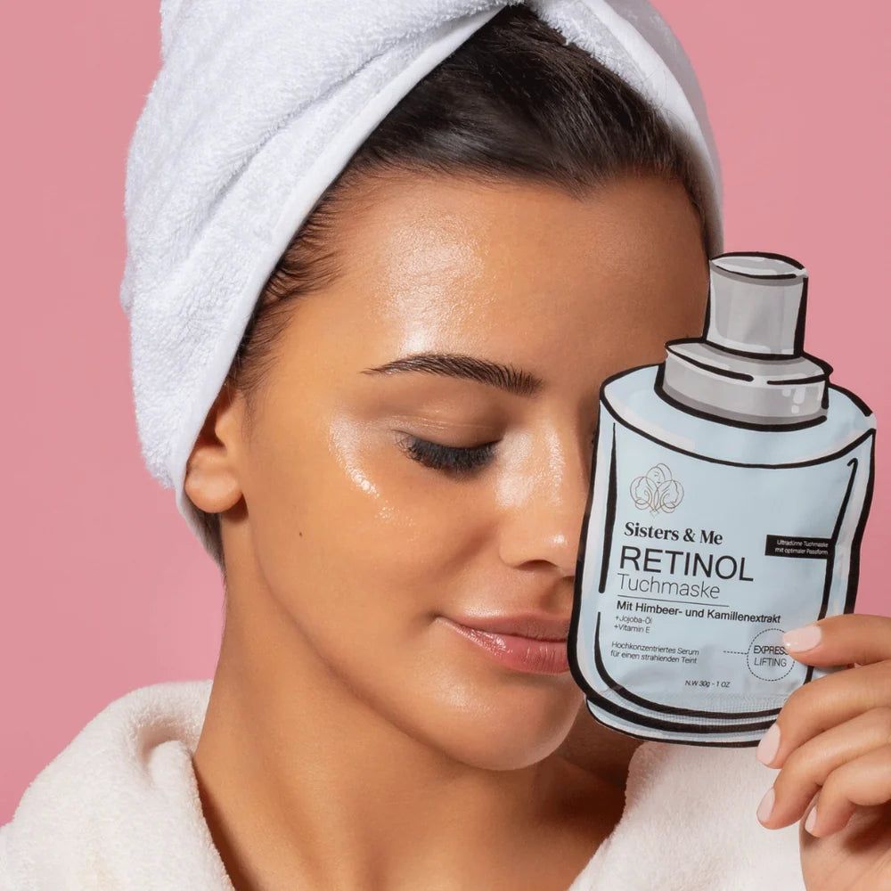 Donna con asciugamano e accappatoio tiene in mano una maschera in tessuto al retinolo. Viso idratato. Sfondo rosa.