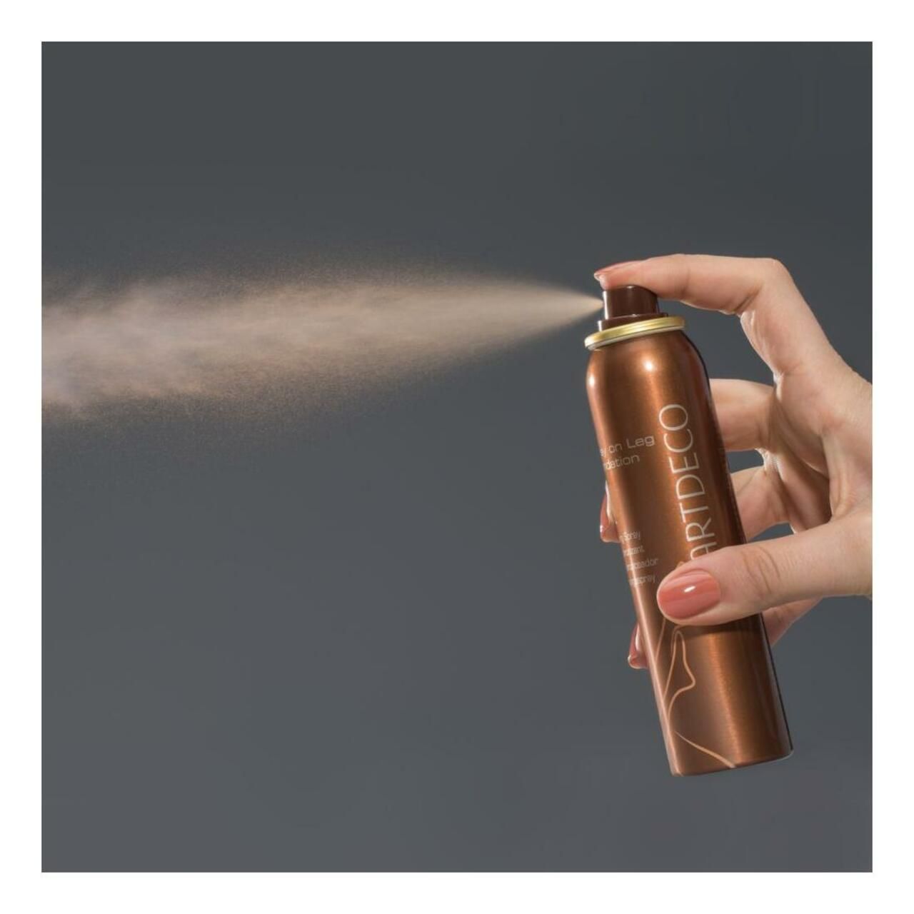 Flacone spray marrone, Artdeco Spray on Leg Foundation, tenuto in mano e che spruzza. Sfondo grigio.