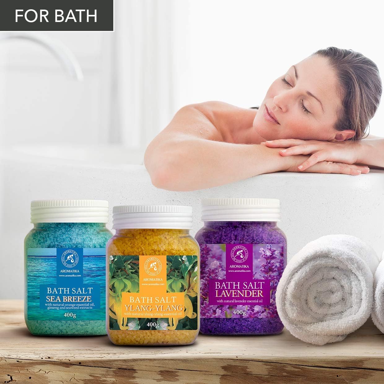 Tre barattoli di sali da bagno accanto a una donna nella vasca. Blu, giallo e viola. Scritte: Sea Breeze, Ylang-Ylang, Lavanda.