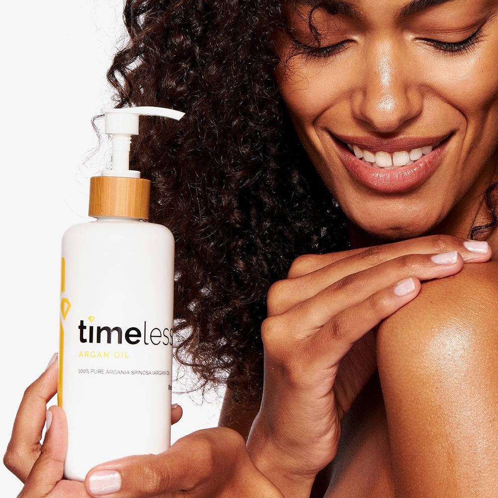 Donna tiene flacone con pompa. Scritta: timeless, Argan Oil. Striscia gialla con logo diamante. Donna tocca la spalla.