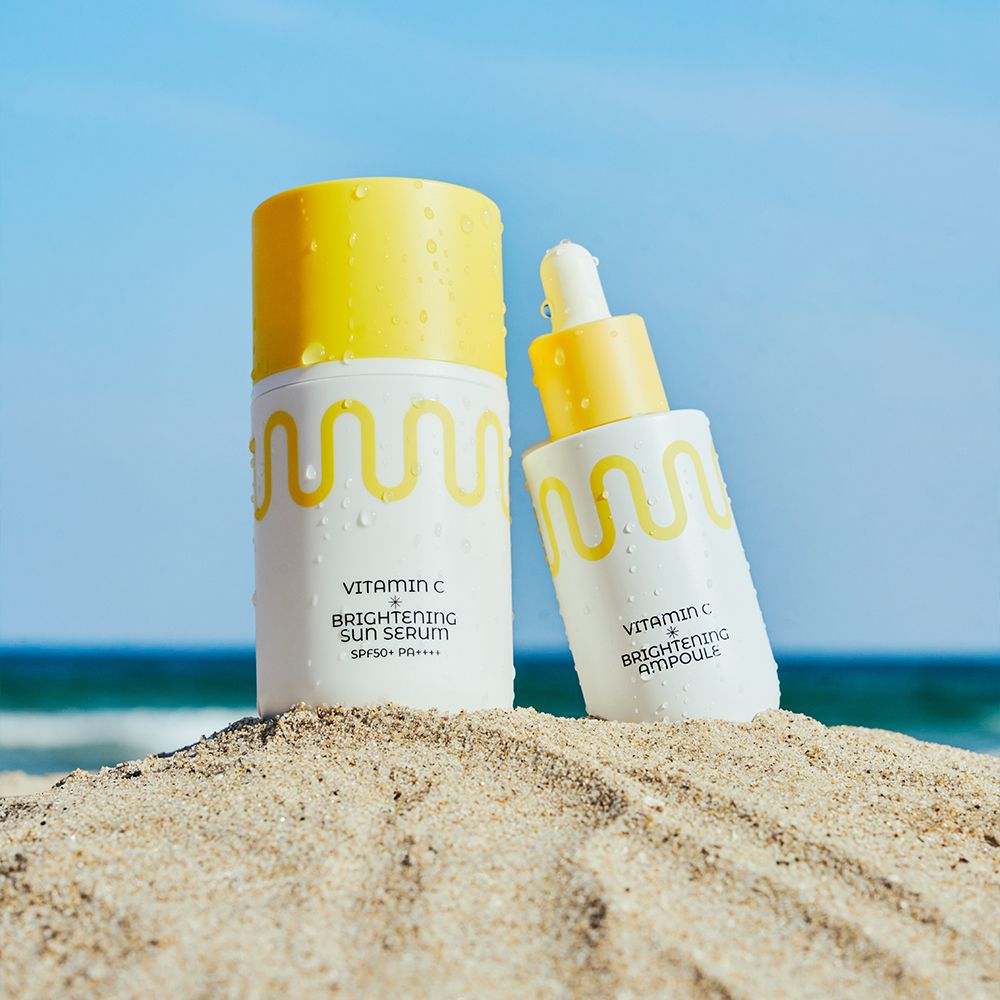 Flacone bianco con tappo giallo e motivo a onde, accanto a un flacone contagocce. Sulla spiaggia.