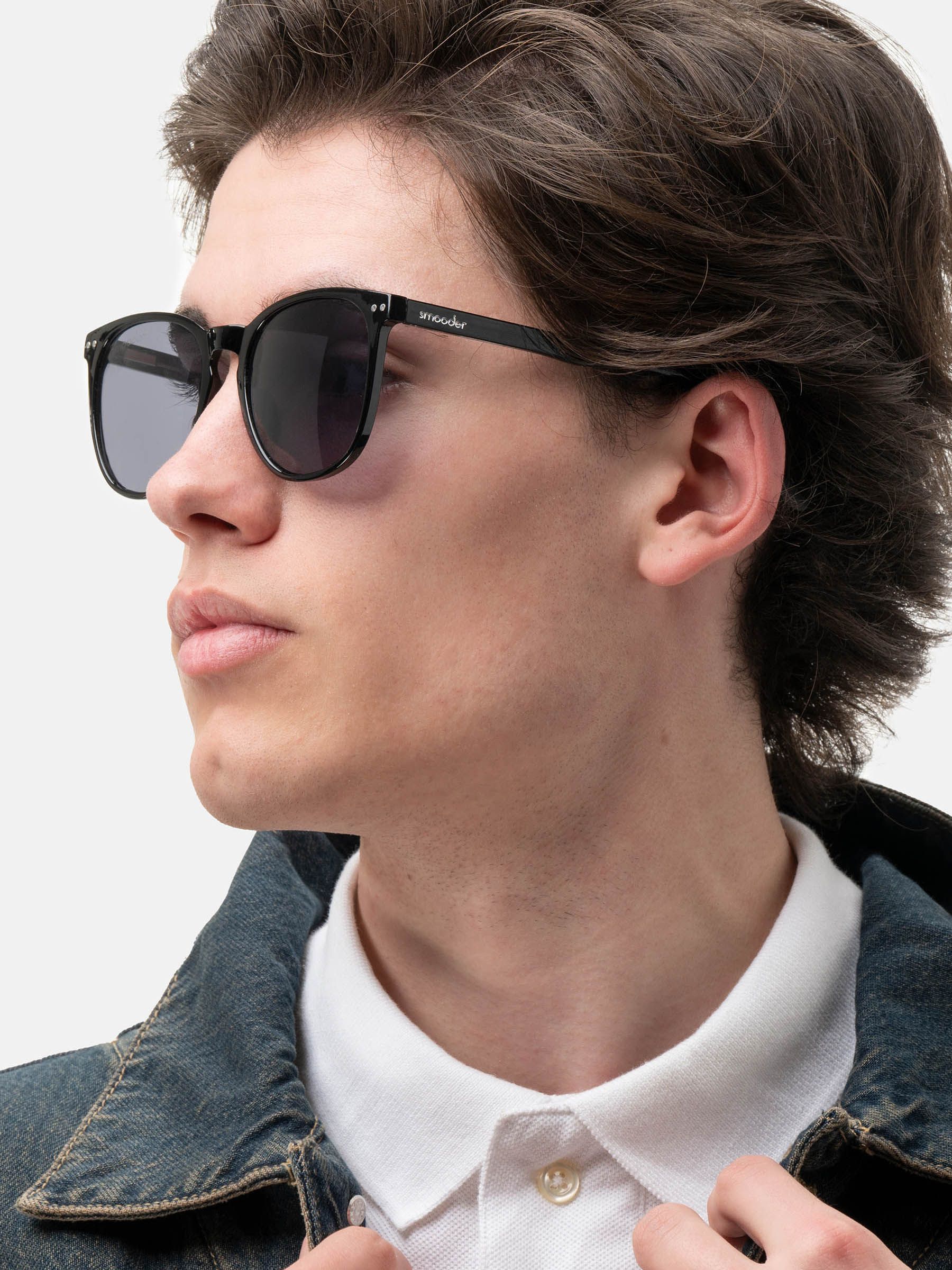 Uomo con occhiali da sole neri con lenti rotonde. Il nome del marchio è visibile sull'asta. Montatura nera.