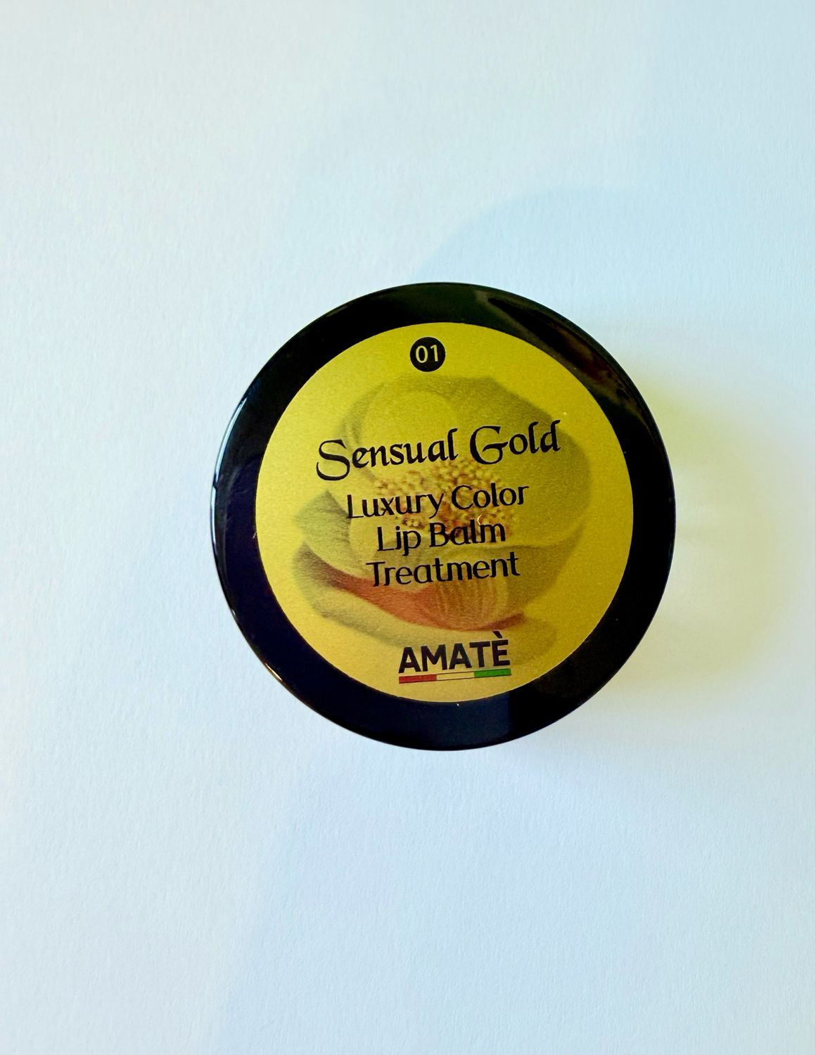 Vista dall'alto di un vasetto di Sensual Gold Lip Balm Treatment. Coperchio nero, adesivo dorato con informazioni e logo AMATÈ.