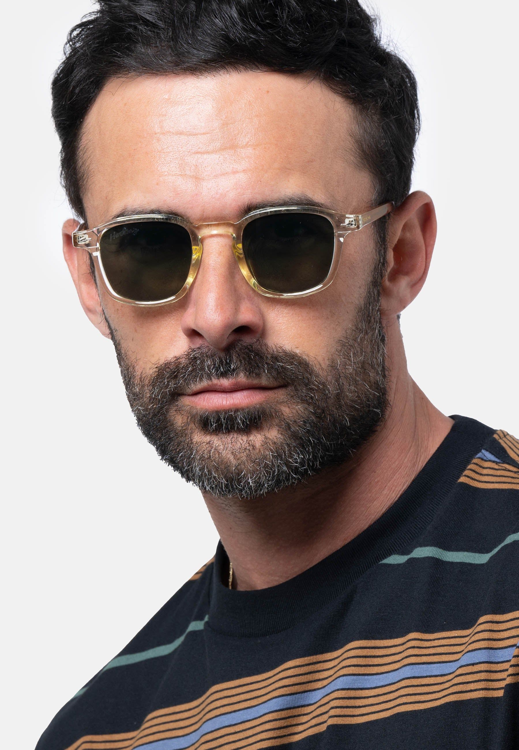 Uomo con occhiali da sole con montatura trasparente e lenti verdi. Il nome del marchio 'Herling' è visibile sulla lente sinistra.