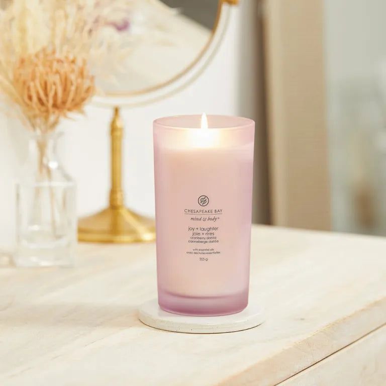 Candela accesa in vetro rosa. Scritta: Chesapeake Bay mind & body, joy + laughter, Cranberry Dahlia. Su sottobicchiere.
