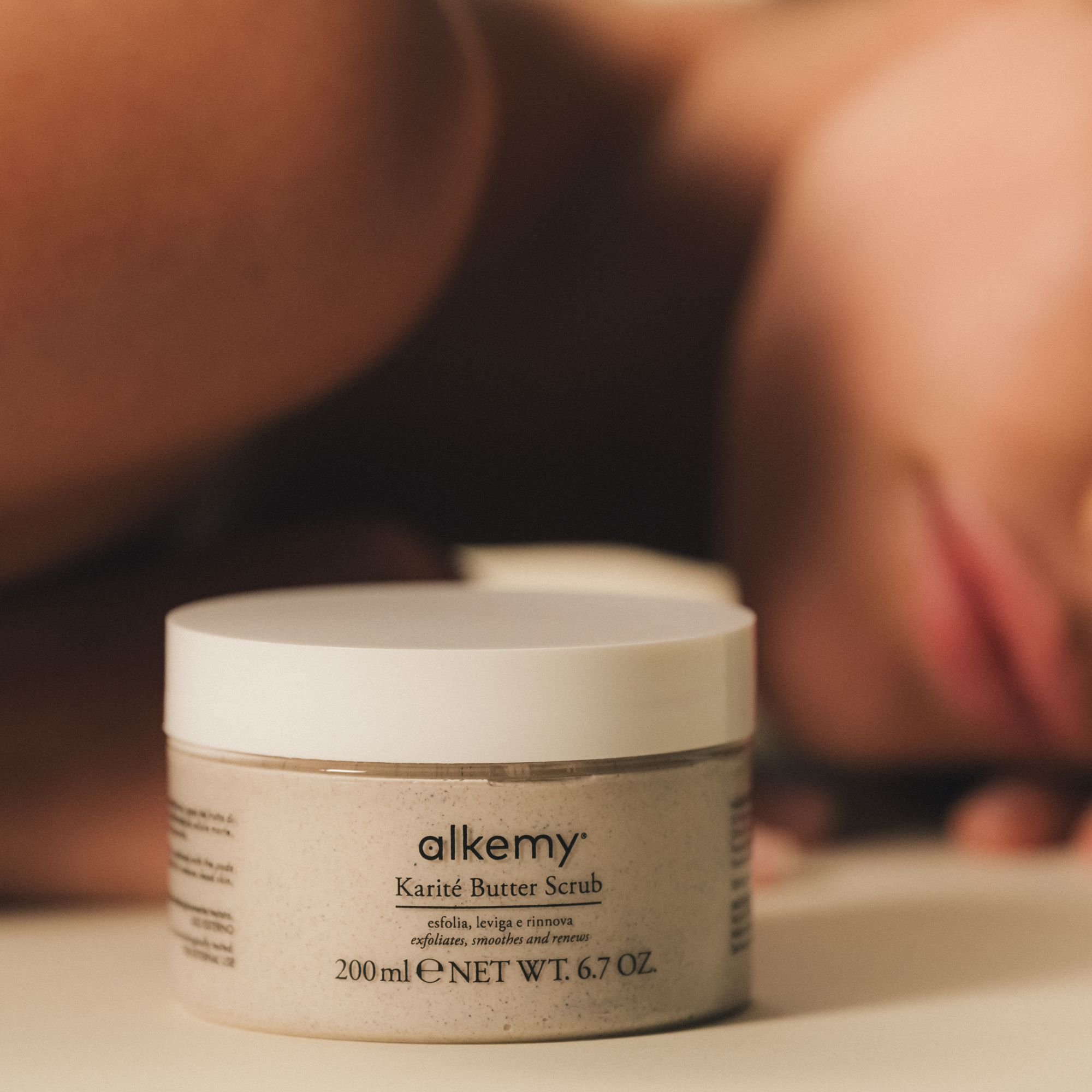 Alkemy Karité Butter Scrub esfoliante corpo in barattolo, accanto a una persona sdraiata.