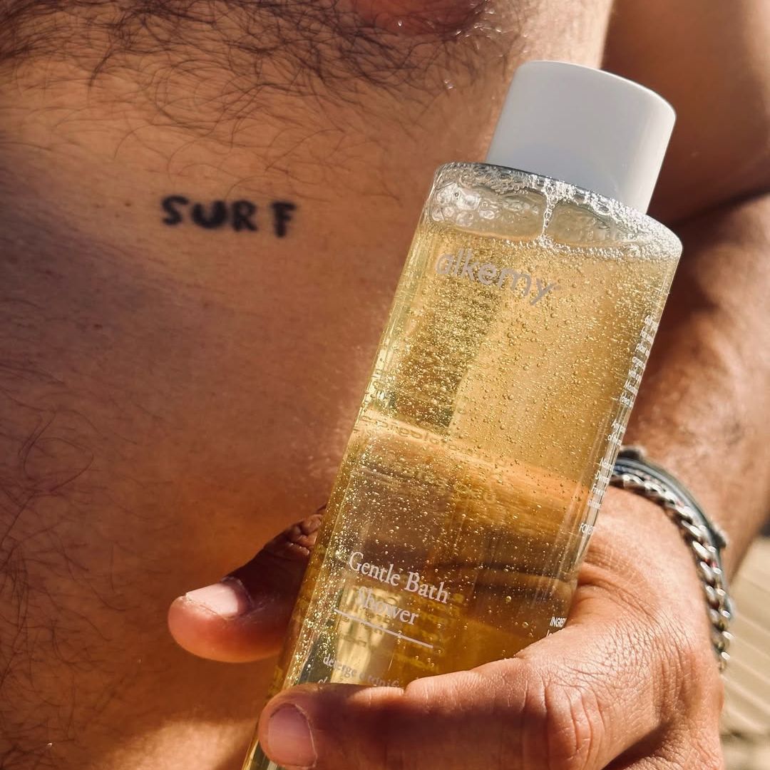 Bagnoschiuma Alkemy in flacone trasparente, tenuto da una mano su pelle tatuata.