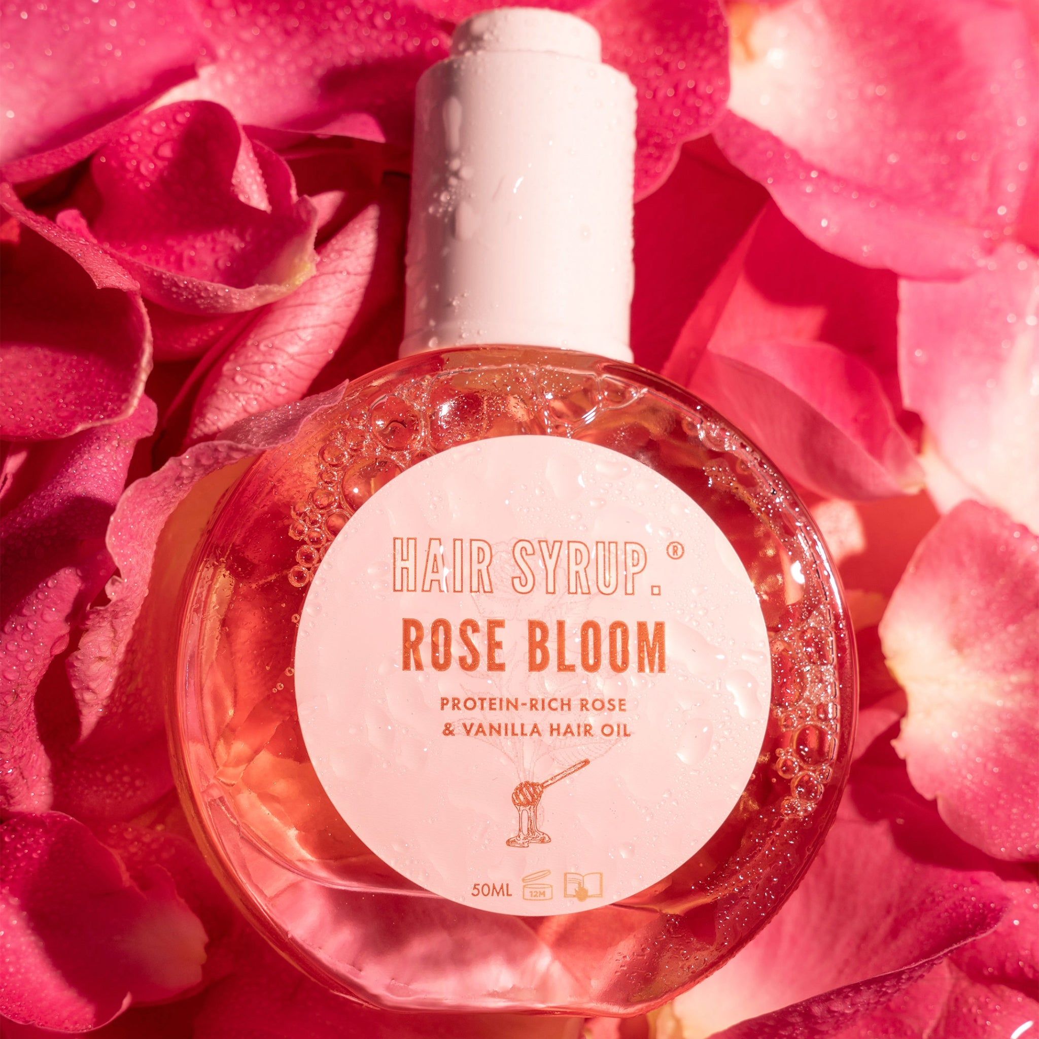 Flacone rotondo con tappo bianco, circondato da petali di rosa. Scritta: Hair Syrup Rose Bloom. Olio per capelli rosa e vaniglia ricco di proteine.