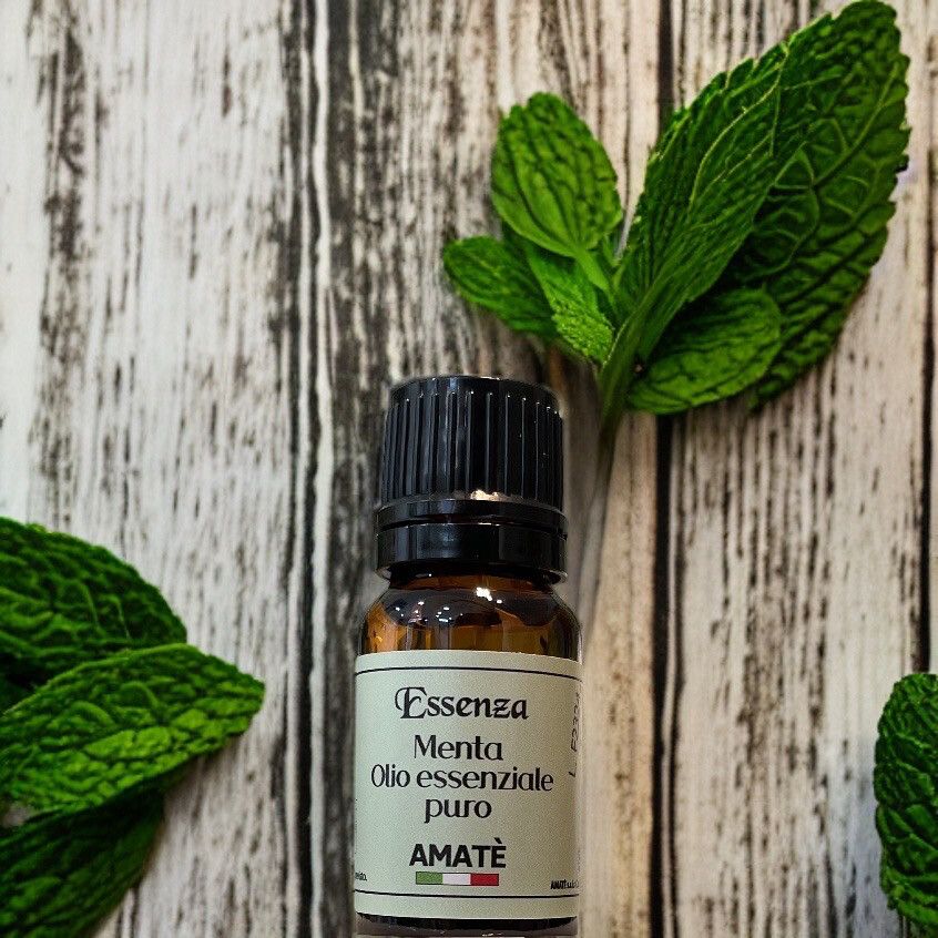 Flacone di vetro marrone con tappo nero, circondato da foglie di menta. Etichetta: Essenza Menta Olio essenziale puro. Marchio: AMATÈ.