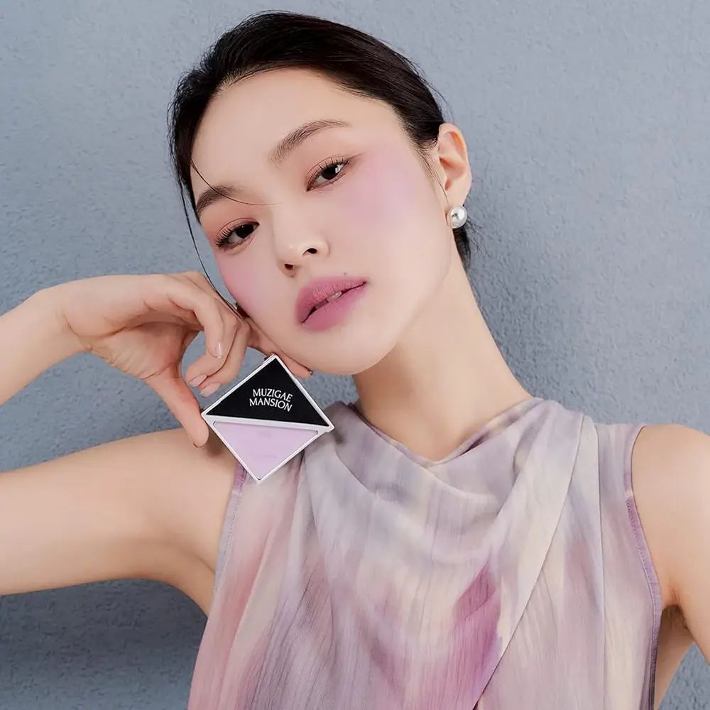 Persona che tiene un fard in forma di diamante. Marchio MUZIGAE MANSION visibile. Tonalità rosa. Trucco sul viso.