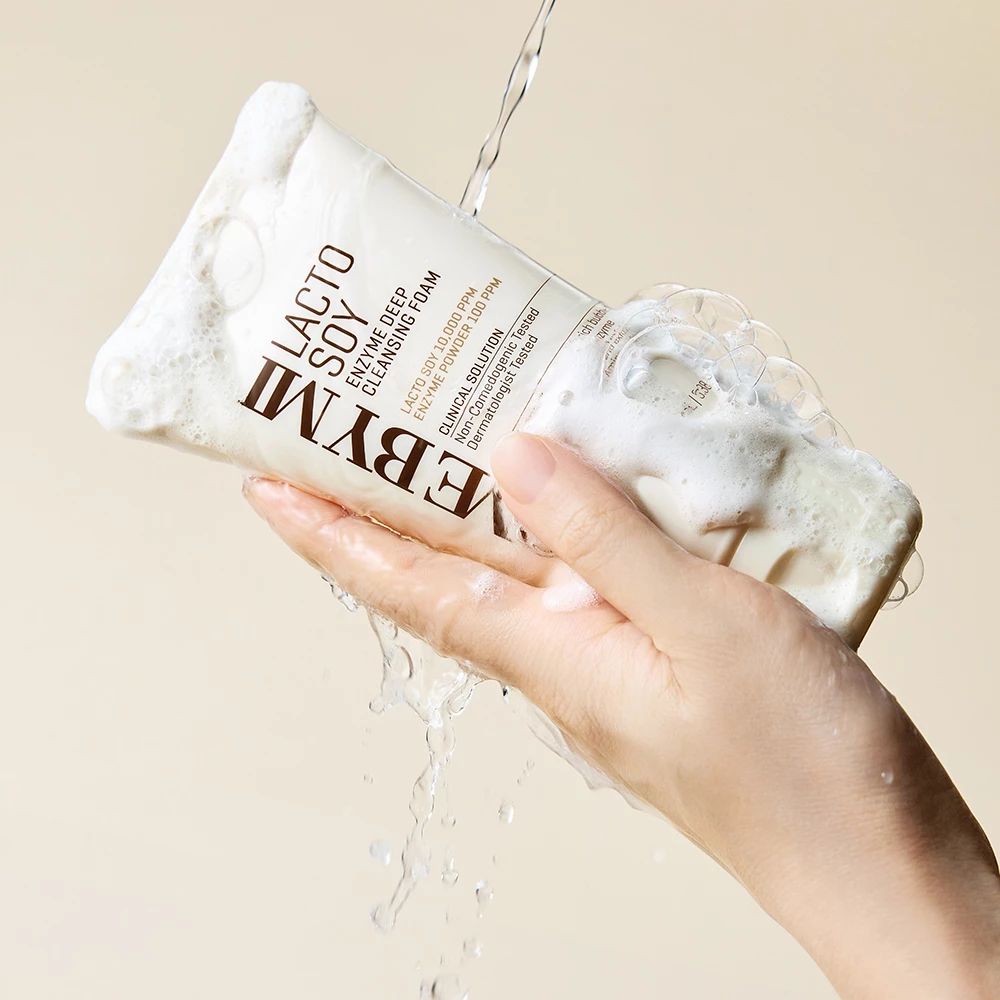 Una mano tiene un tubetto di schiuma detergente bianca. L'acqua scorre sul tubetto e sulla schiuma. Sul tubetto c'è scritto "Lacto Soy Enzyme Deep Cleansing Foam".