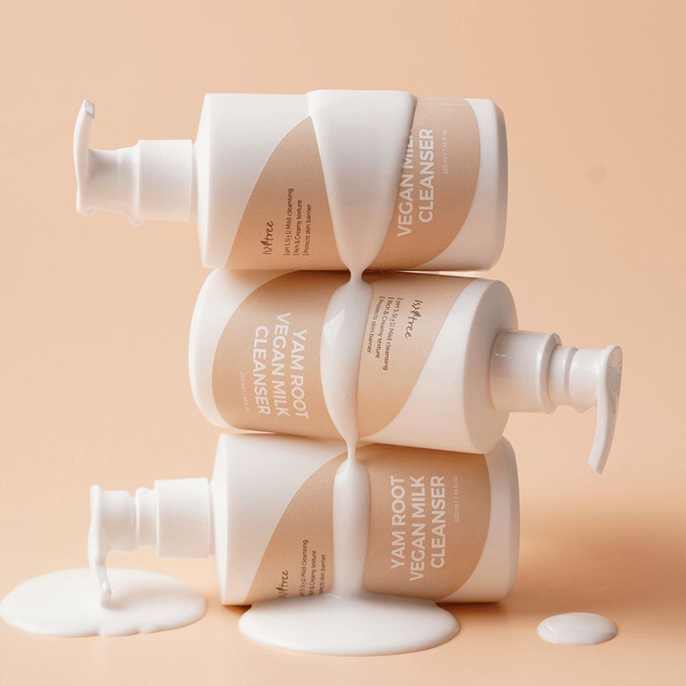 Tre flaconi di detergente viso, bianchi con etichetta beige. Nome del prodotto e marchio visibili. Liquido che cola.