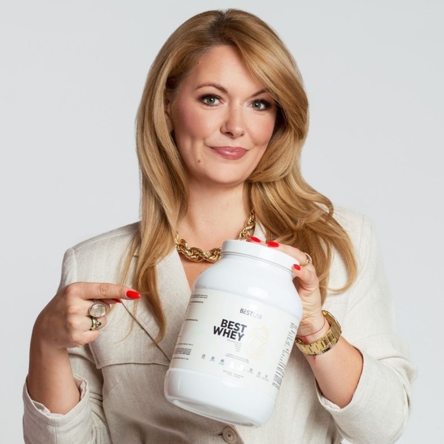 Donna che tiene in mano un barattolo di proteine Best Lab Best Whey bianco. Logo e scritta "BEST WHEY" sul davanti. Lo indica.